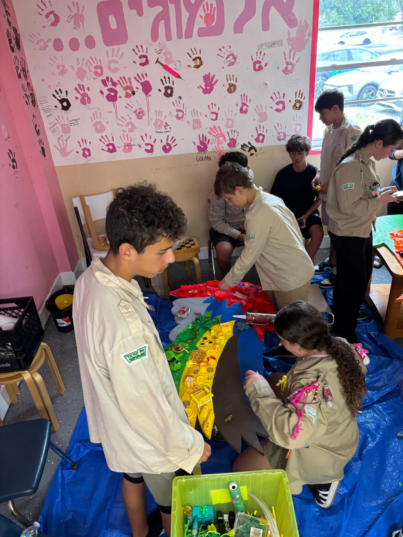 חניכים במדים עובדים בחדר עם קיר טביעות יד ורודות